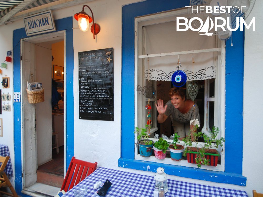 Friendly restaurant owner waving goodbye to guests from the window of her small Bodrum eatery.